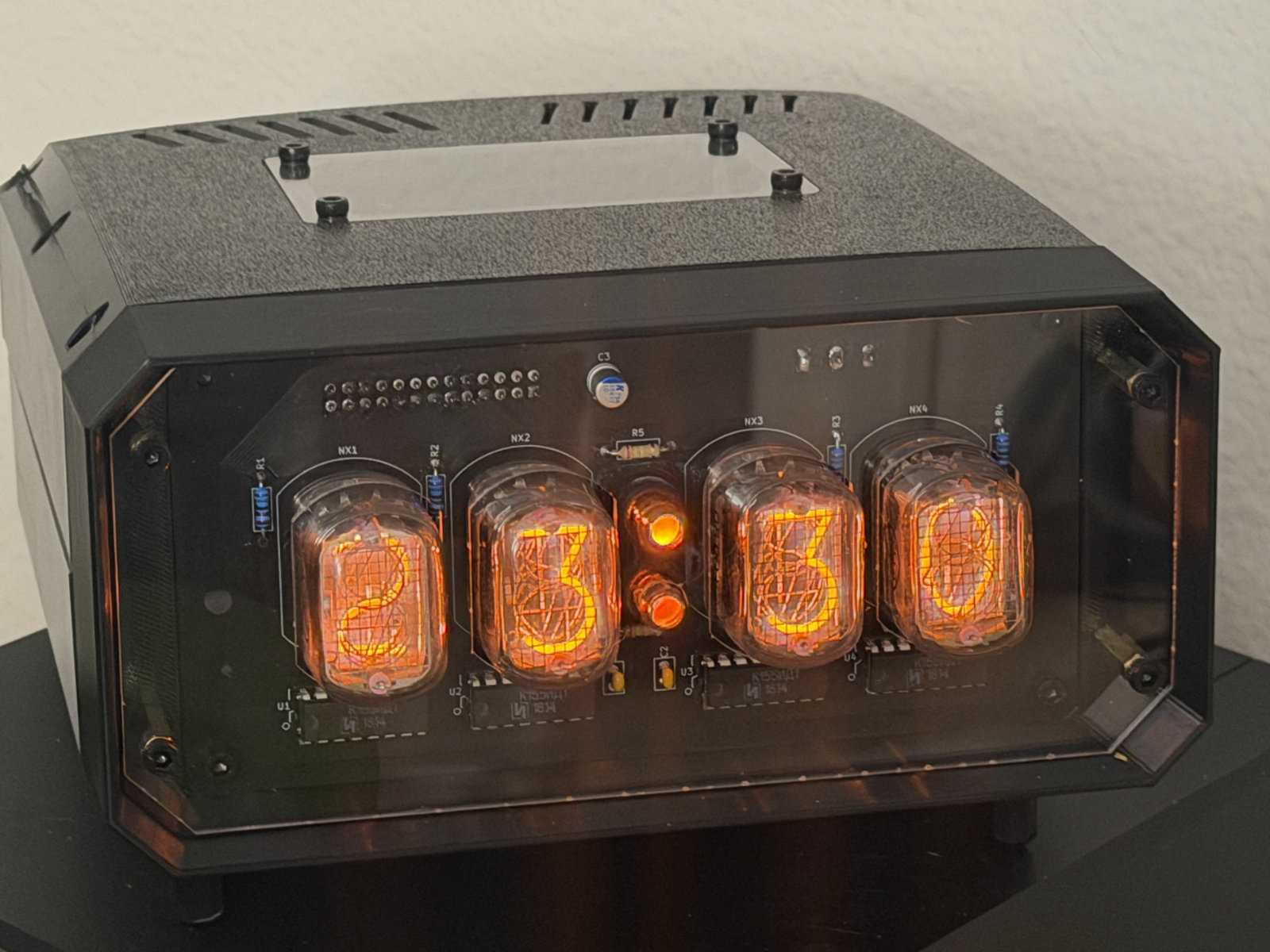 Four nixie tubes glowing orange behind a acrylic panel, reading 23:30, with a neon pair of dots between the hour and minute pairs. The exposed board behind them is silkscreened NX1 to NX4 for the tubes and R1 to R5 for the resistors. A black 3D printed case with a vented top encloses it.