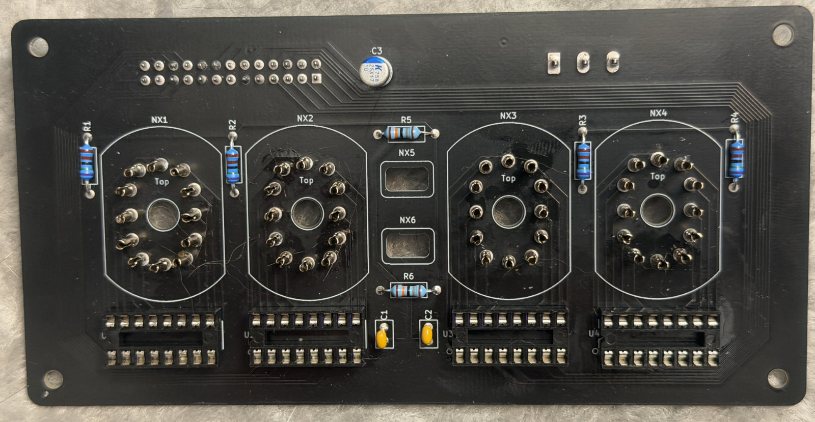 A bare black circuit board with four eleven-pin tube sockets silkscreened NX1 to NX4, two rectangular pads for the colon tubes NX5 and NX6, six axial resistors, an electrolytic capacitor at C3, and four empty DIP sockets along the bottom edge.